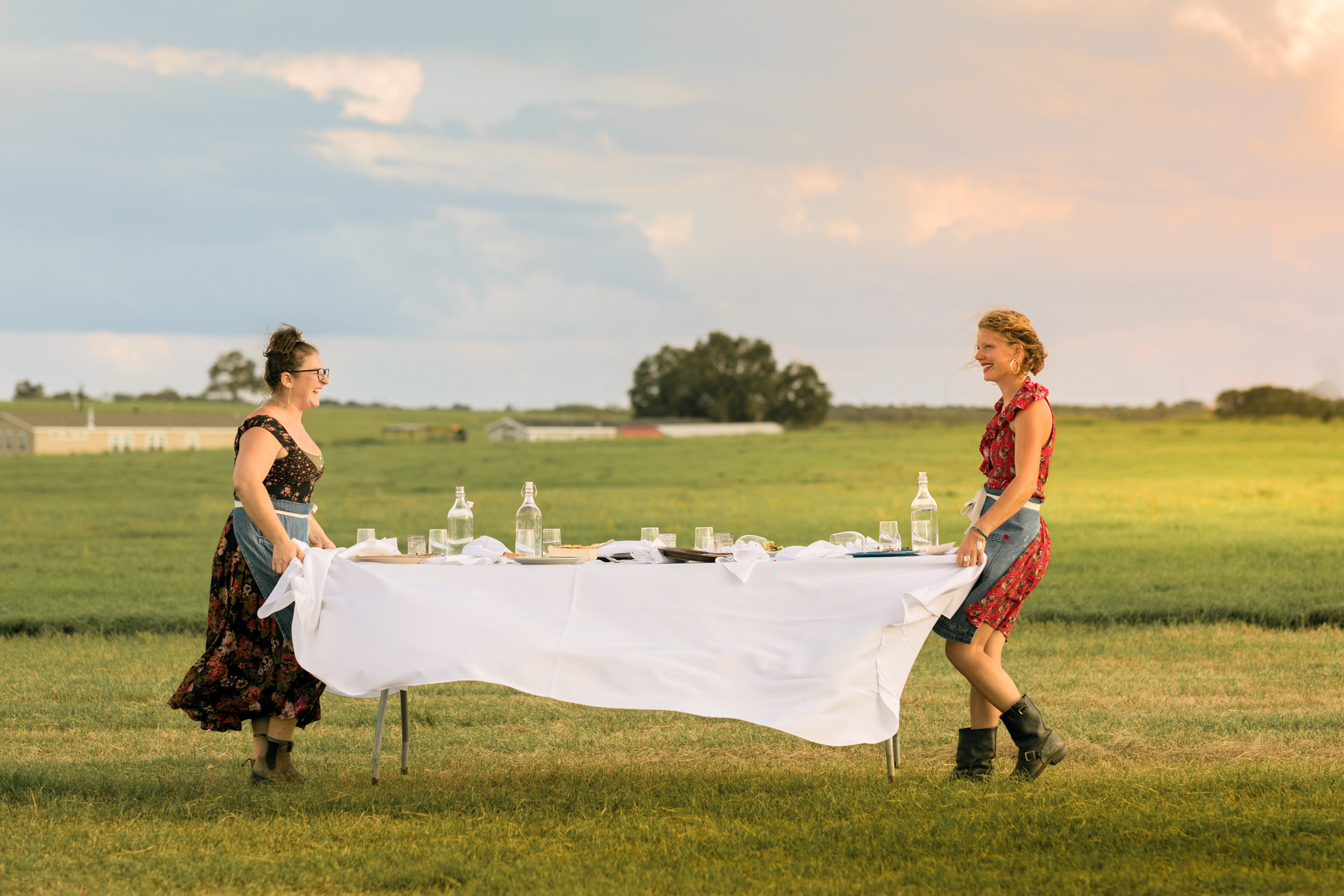two woman carry table through large grassy field on farm