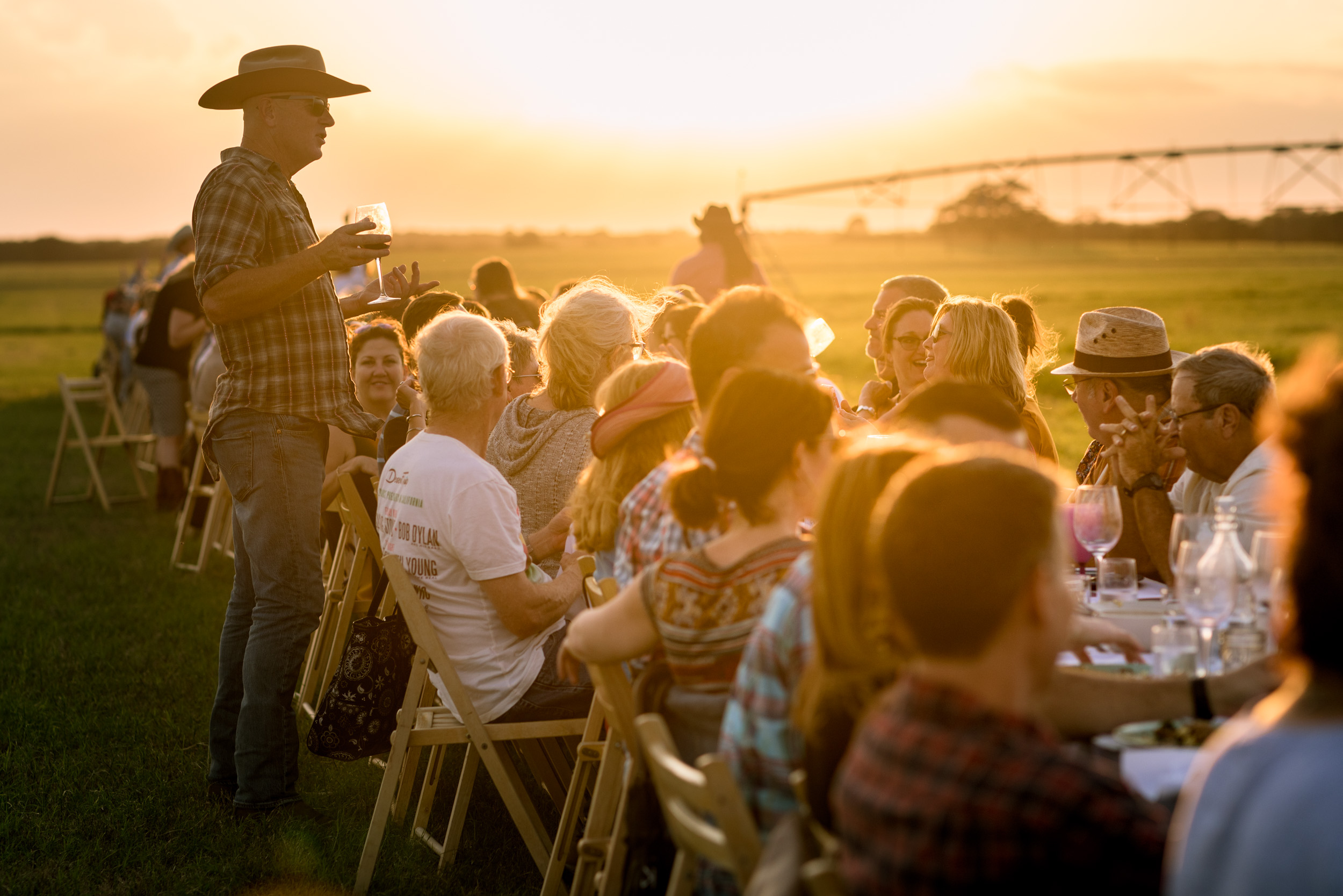 long table filled with people eating dinner on farm outside at sunset