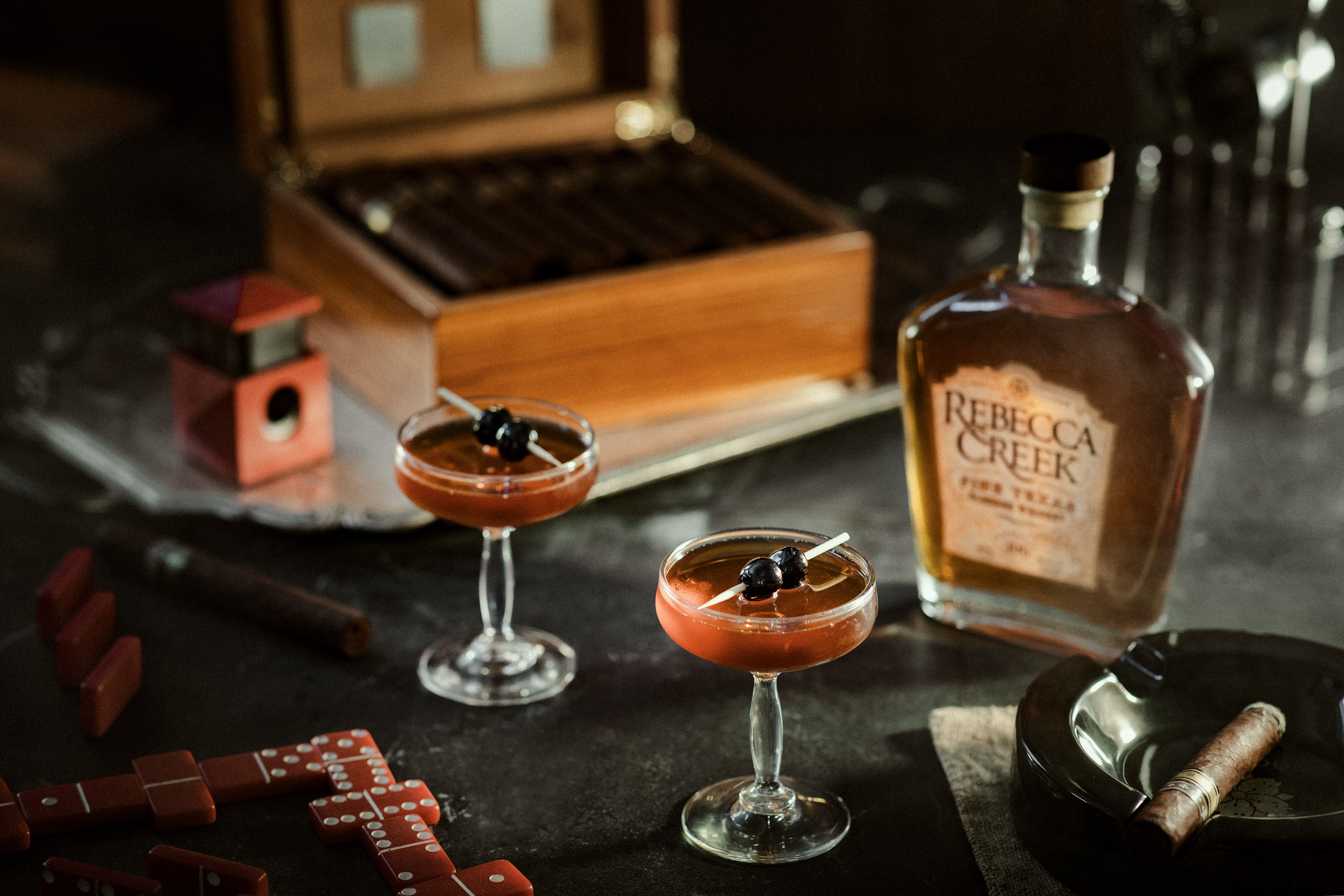 two cocktails in coupe glasses with cigar box and rebecca creek bottle