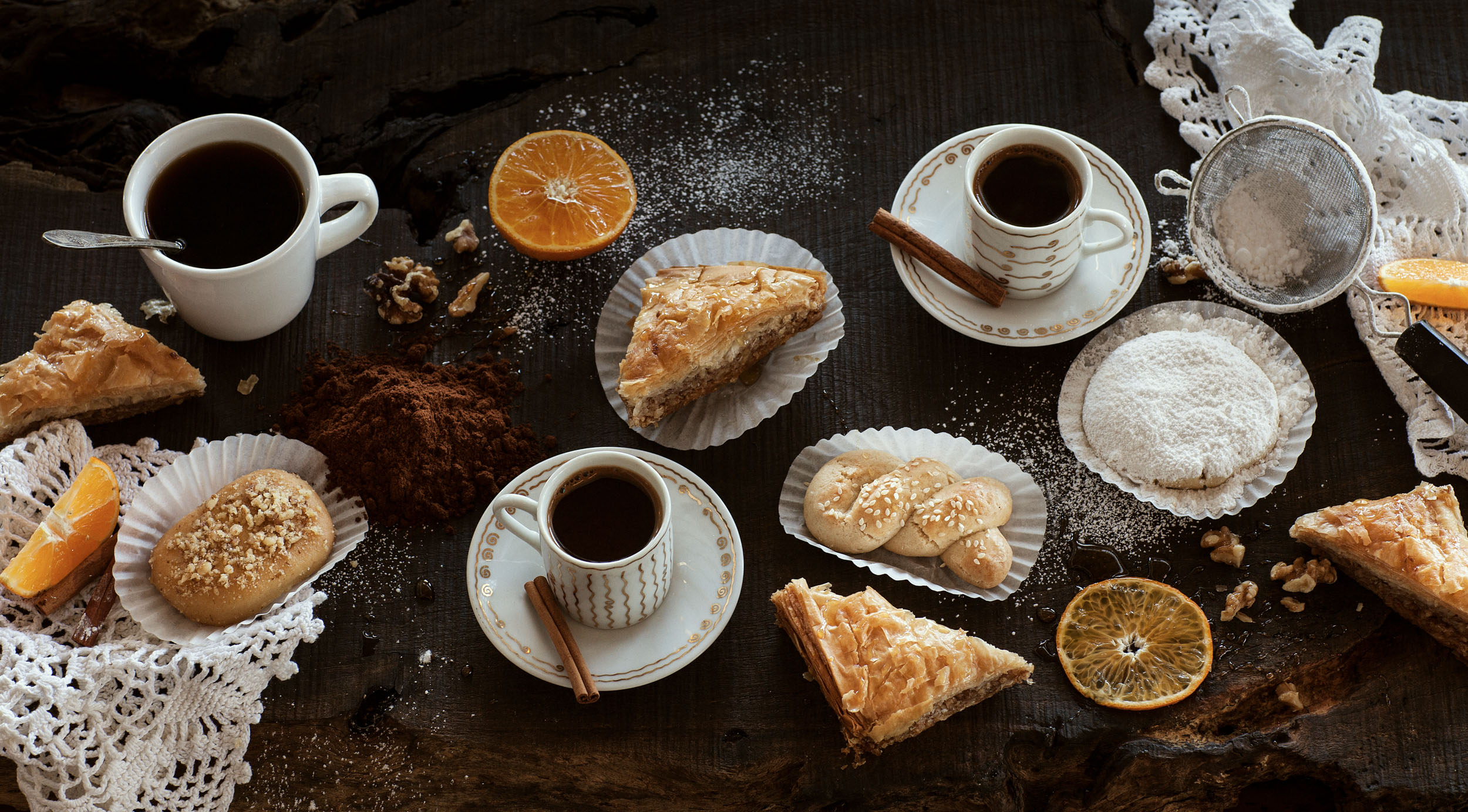 large spread of greek desserts and coffee on dark surface
