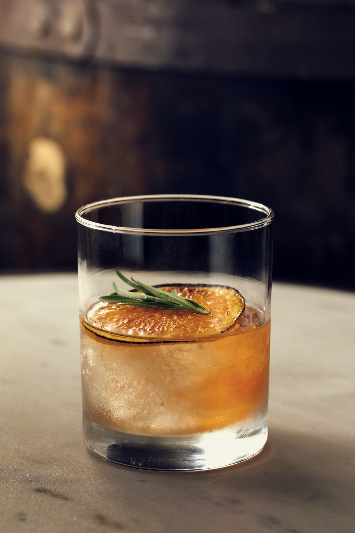 whiskey cocktail with dried orange slice and rosemary garnish on table with barrel