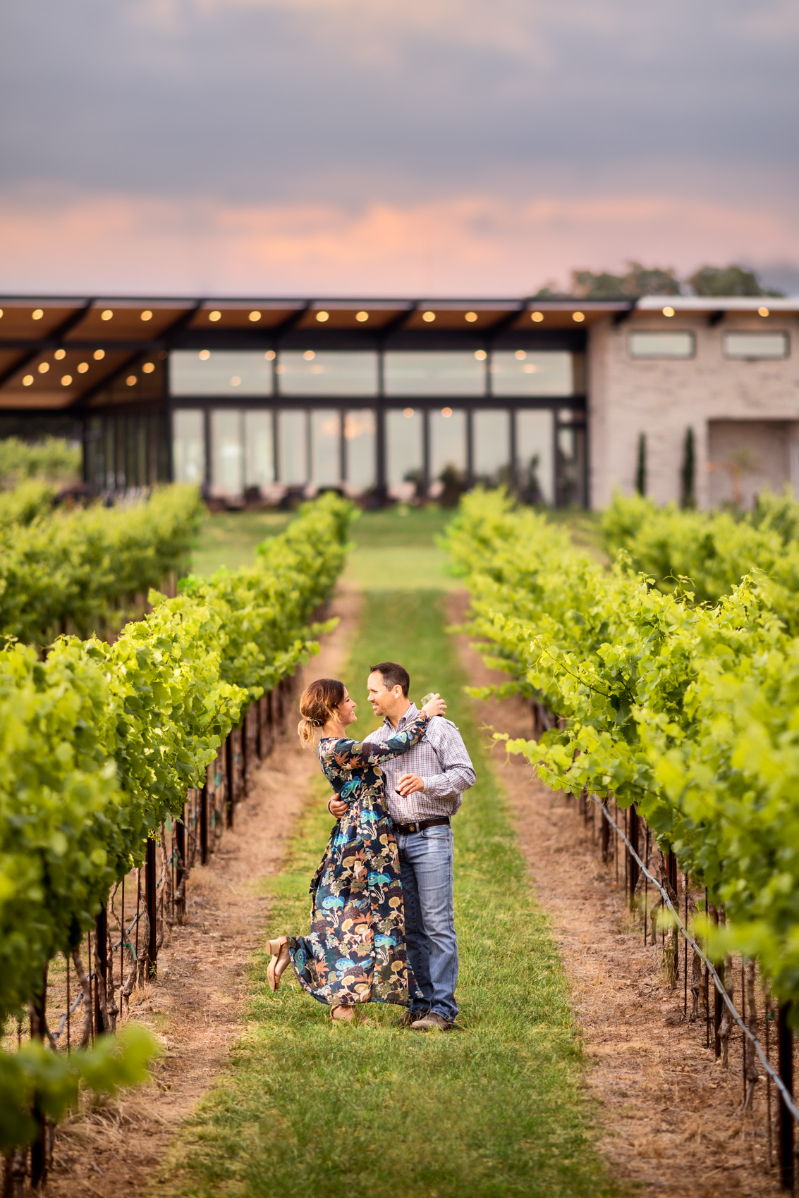 couple embrace in vineyard with winery in background