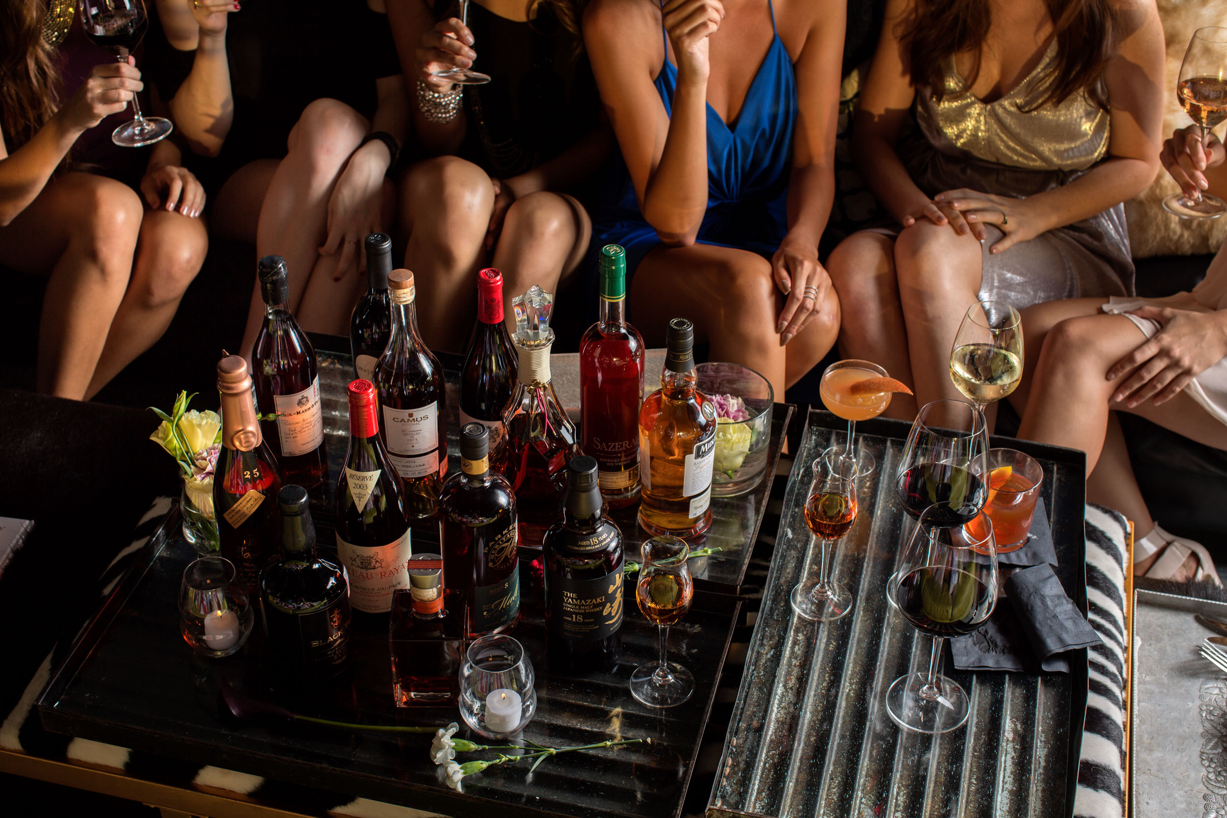 table of spirit bottles and cocktails with women sitting on couch