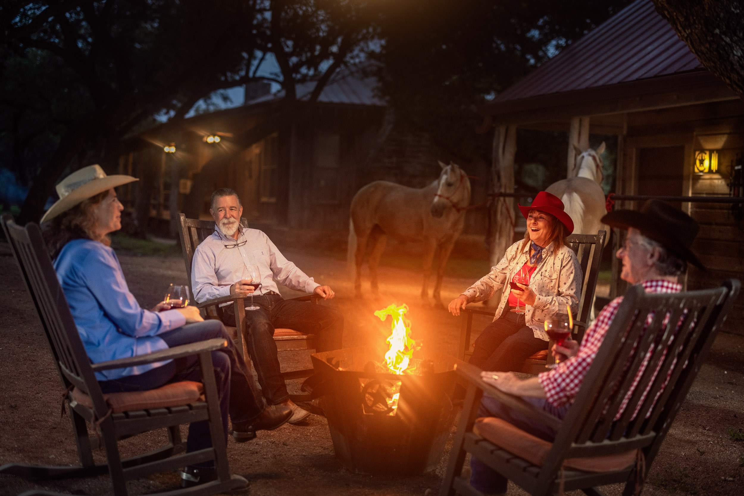 four people sitting around campfire at night with horses in background